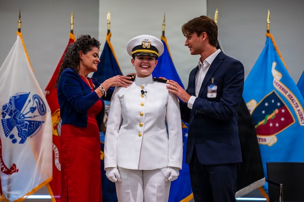 (260710-N-AJ782-1143) FALLS CHURCH, Va. U.S. Navy Ensign Jenna Hosea Baker, center, a nurse assigned to Walter Reed National Military Medical Center, receives her shoulder boards from her mother, Deanna McAlister Hosea, left, and her husband, Andrew Baker, right, as part of a joint ceremony featuring the retirement of her father at Defense Health Headquarters in Falls Church, Virginia, July 10, 2026. The retirement of Capt. Lonnie S. Hosea, Bureau of Medicine and Surgery senior health policy advisor, and the commissioning of Baker highlighted a continuing legacy of service for Navy Medicine as Hosea closed out a 30-year career and Baker took her first steps into the Nurse Corps. (U.S. Navy photo by Kevin Ray J. Salvador)