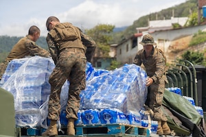 Three Marines in camouflage uniforms remove pallets of water from the back of a military truck.