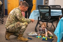 A uniformed service member crouches to engage with a young child at an event with tables, chairs, and attendees visible throughout the room.