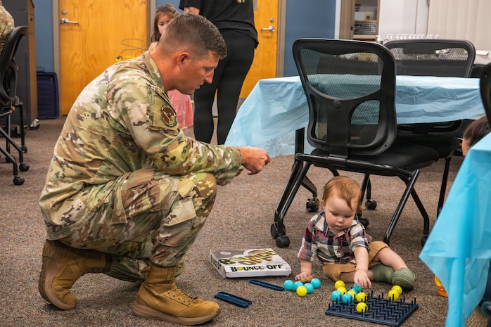 A uniformed service member crouches to engage with a young child at an event with tables, chairs, and attendees visible throughout the room.