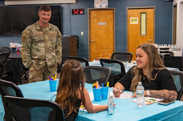 A service member smiles while talking with a woman and child seated at a decorated table.