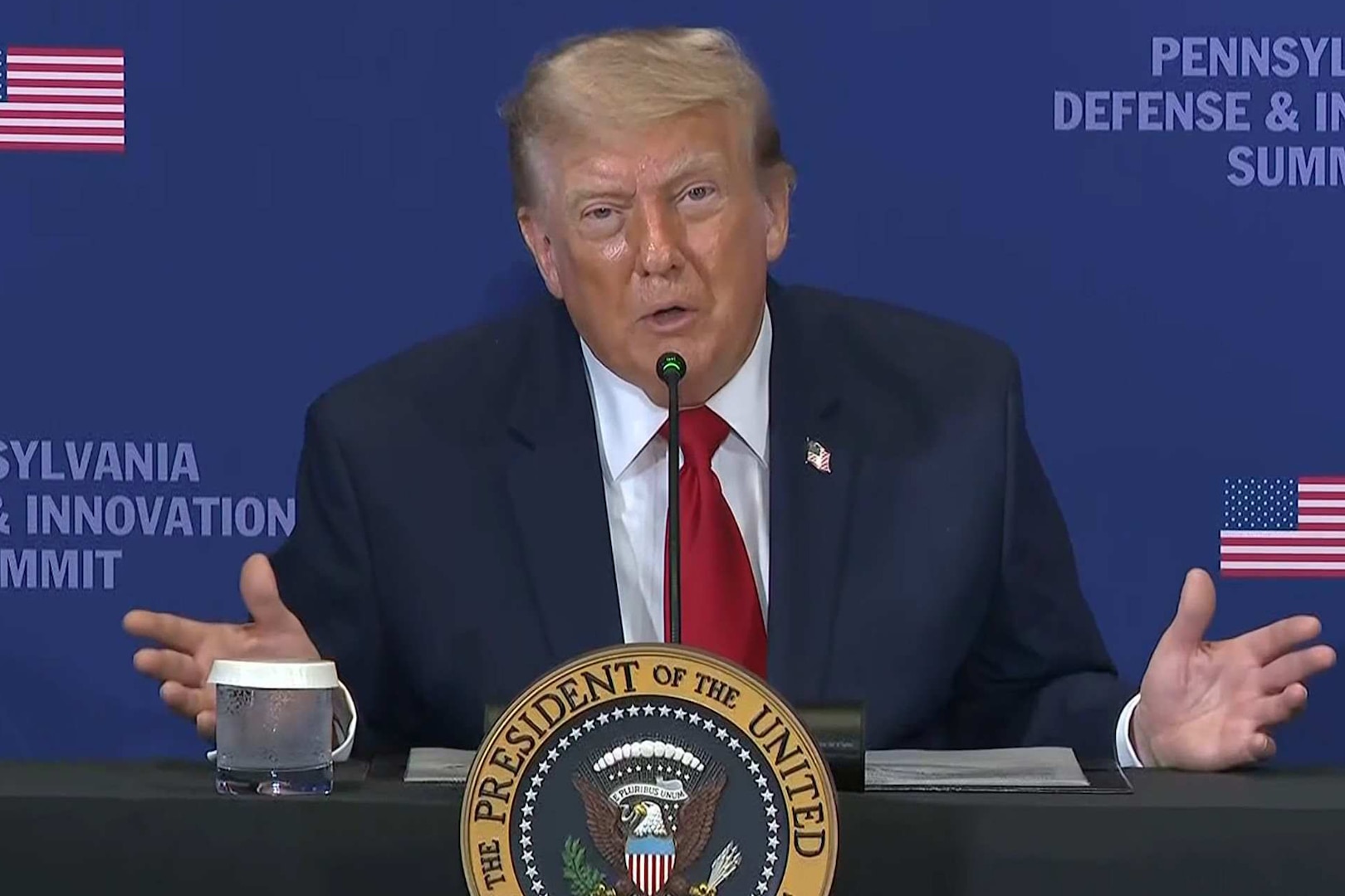 A man in a suit speaks into a microphone while seated at a table that has the presidential seal in front of it.