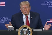 A man in a suit speaks into a microphone while seated at a table that has the presidential seal in front of it.