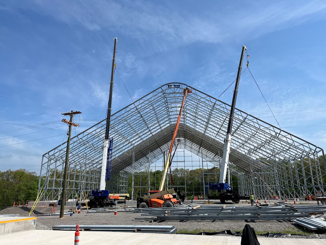 Two large mobile cranes and an orange boom lift are positioned around the steel skeleton of a large, arched building under construction against a blue sky.