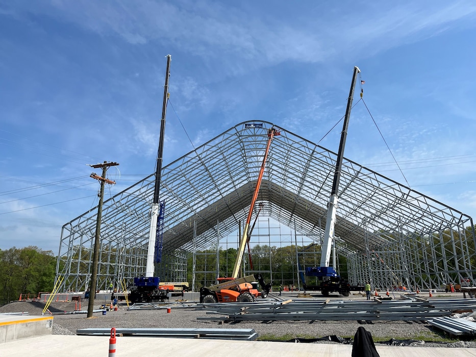 Two large mobile cranes and an orange boom lift are positioned around the steel skeleton of a large, arched building under construction against a blue sky.
