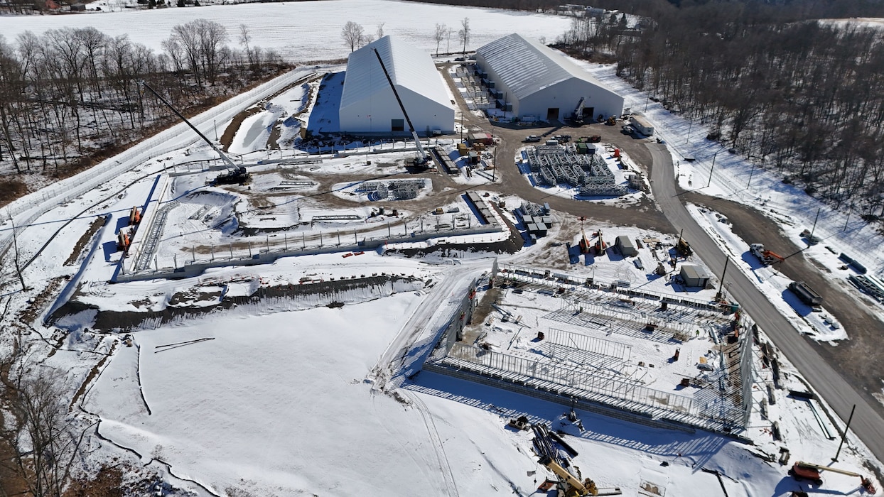 An aerial view of a snowy construction site shows two large white buildings and the foundation work for new structures, with cranes and construction equipment visible.