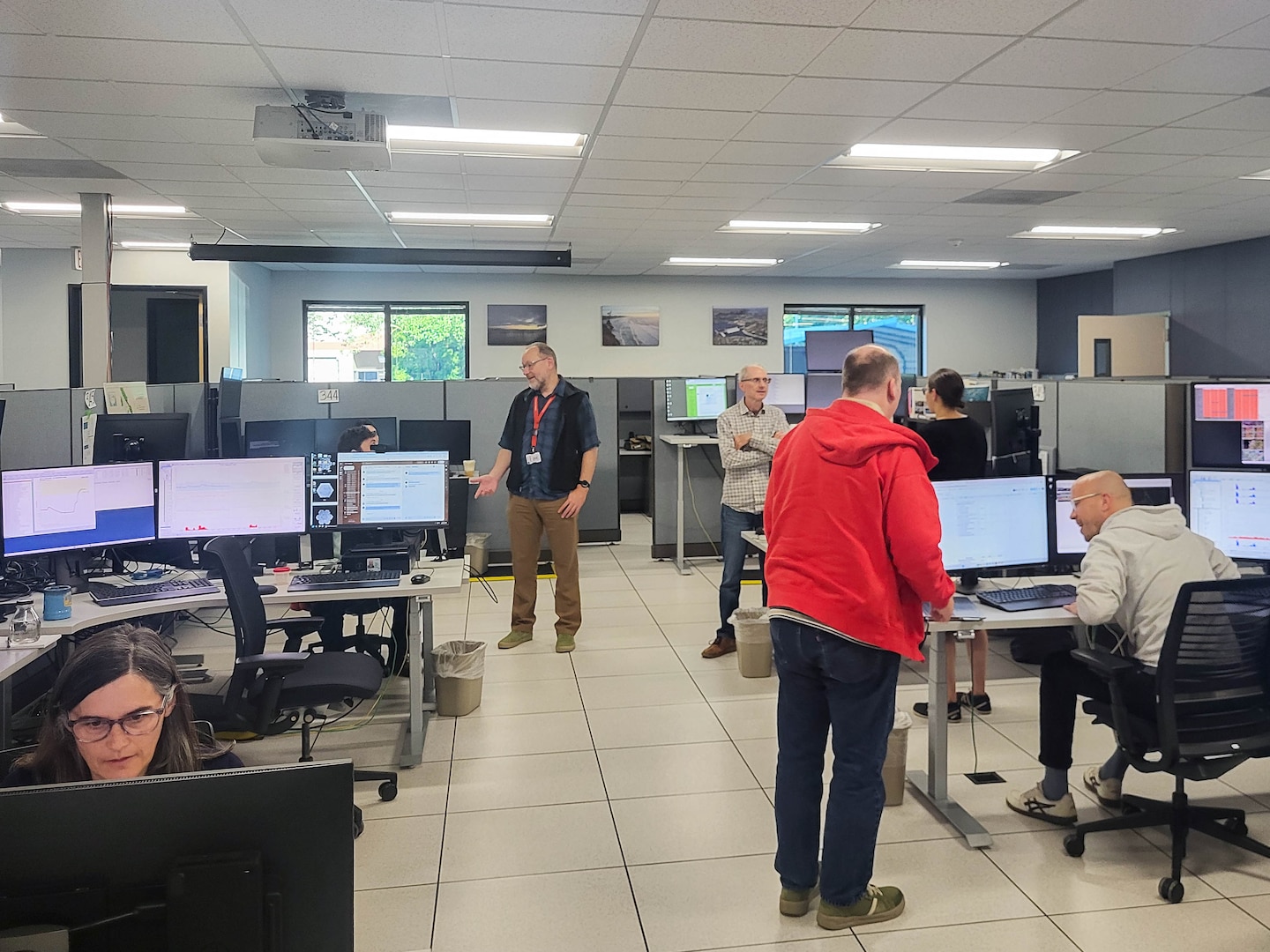 Several people stand and sit in an office space with computers, speaking with each other and looking at the computers.