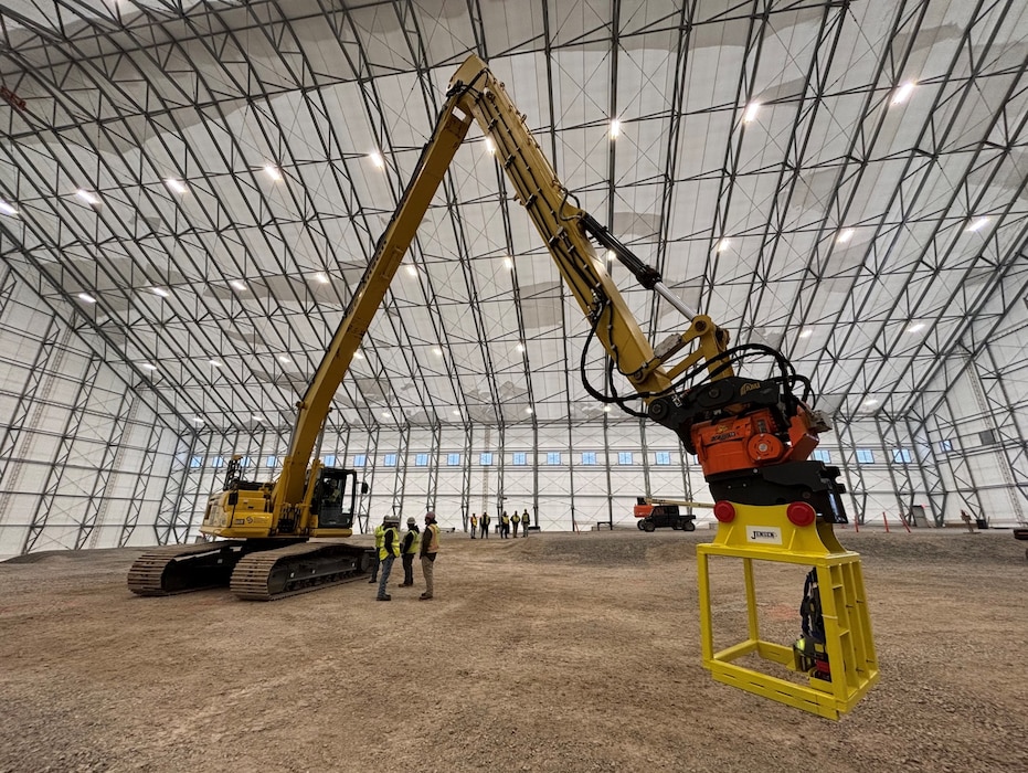 A low-angle shot of a very large yellow excavator with a long arm and a shear attachment inside a cavernous, white, arched structure.