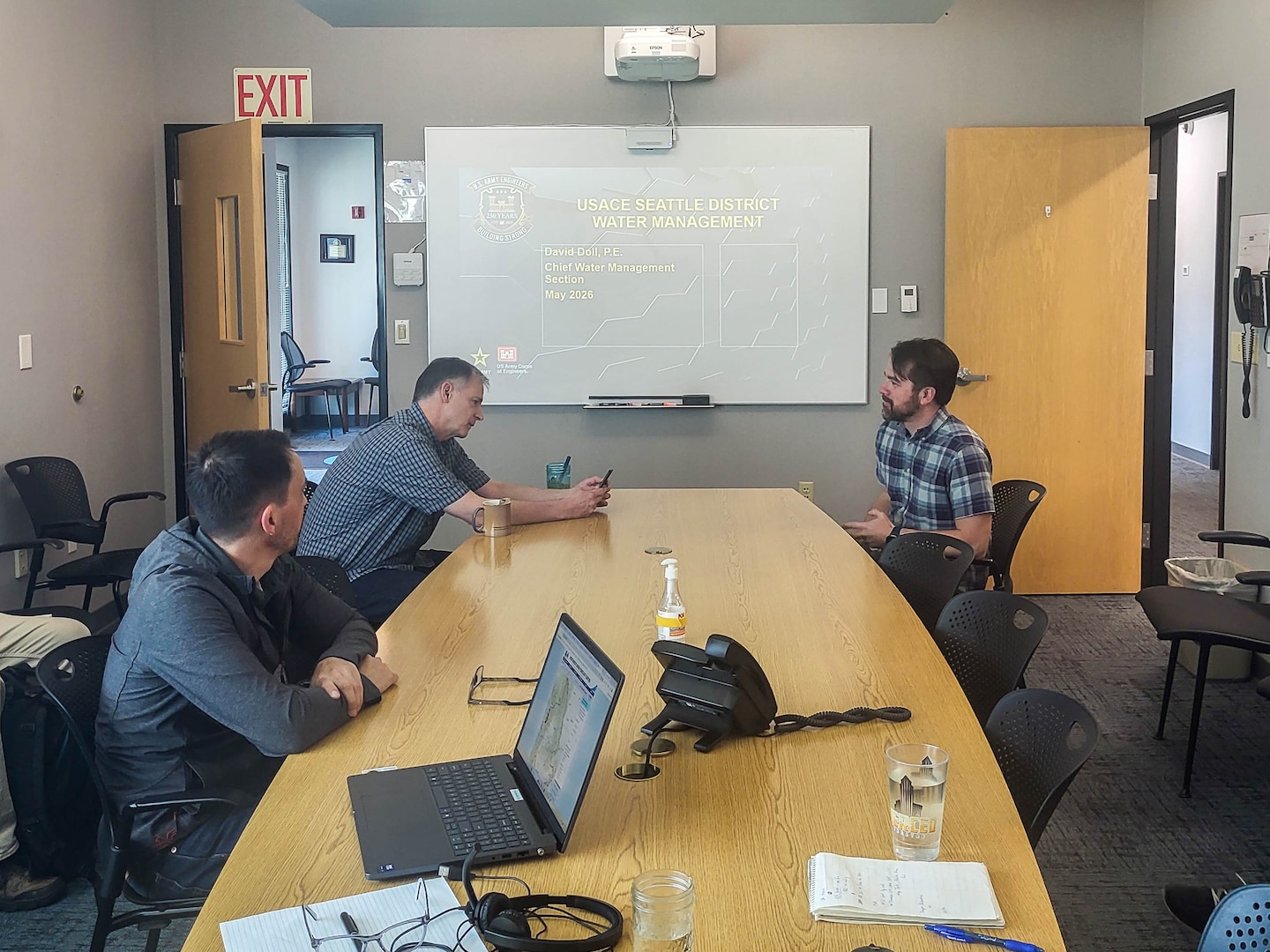 Three people sit at a conference room table, with a slideshow presentation on the screen by them that reads "USACE Seattle District Water Management"