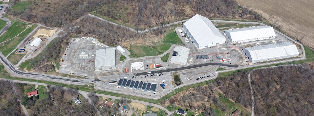 An aerial overview of a sprawling industrial site featuring several large, hangar-like buildings and smaller structures, surrounded by bare trees and green grass.