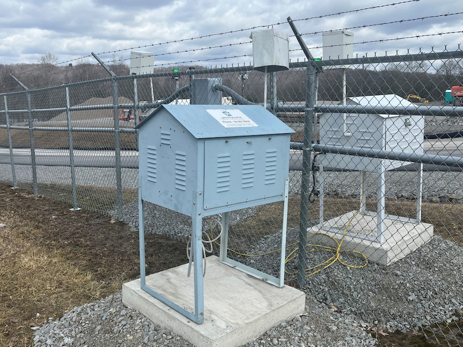 A gray, house-shaped metal box sits on a concrete pad next to a chain-link fence at an industrial site.