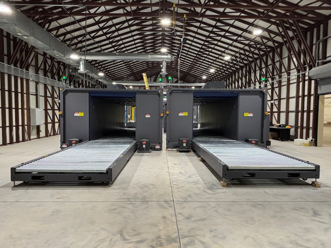 Two large, gray industrial scanning machines with roller conveyors sit parallel to each other inside a large warehouse with an arched metal ceiling.