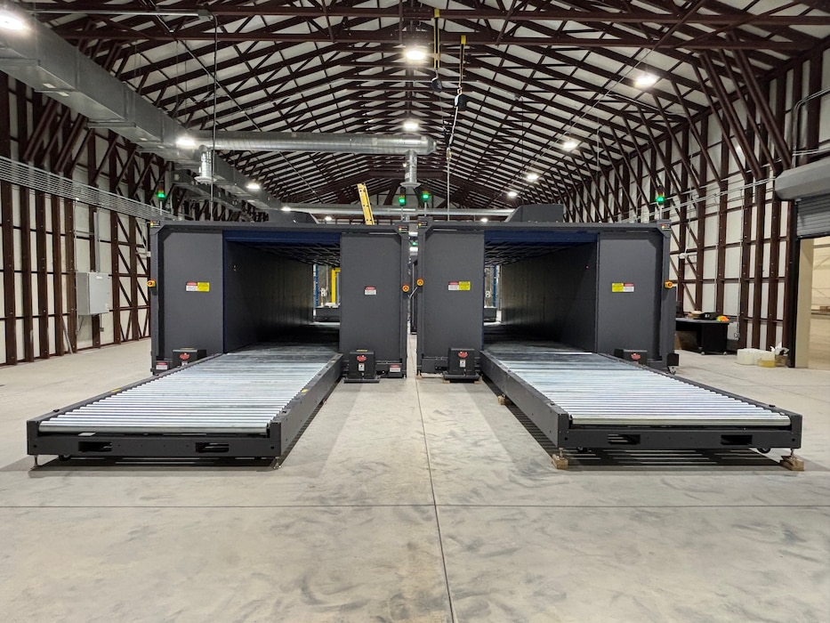 Two large, gray industrial scanning machines with roller conveyors sit parallel to each other inside a large warehouse with an arched metal ceiling.