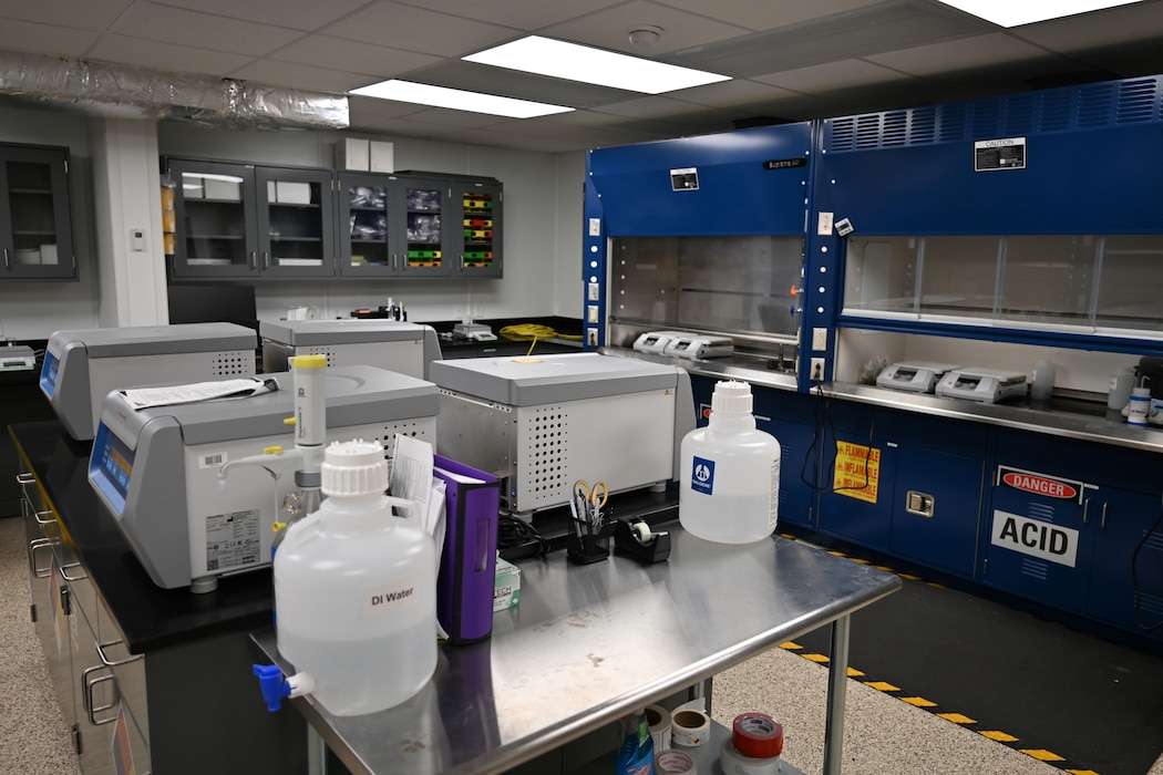 An interior view of a science laboratory with stainless steel counters, blue chemical fume hoods, and various pieces of equipment. A "DANGER ACID" sign is visible on a cabinet.