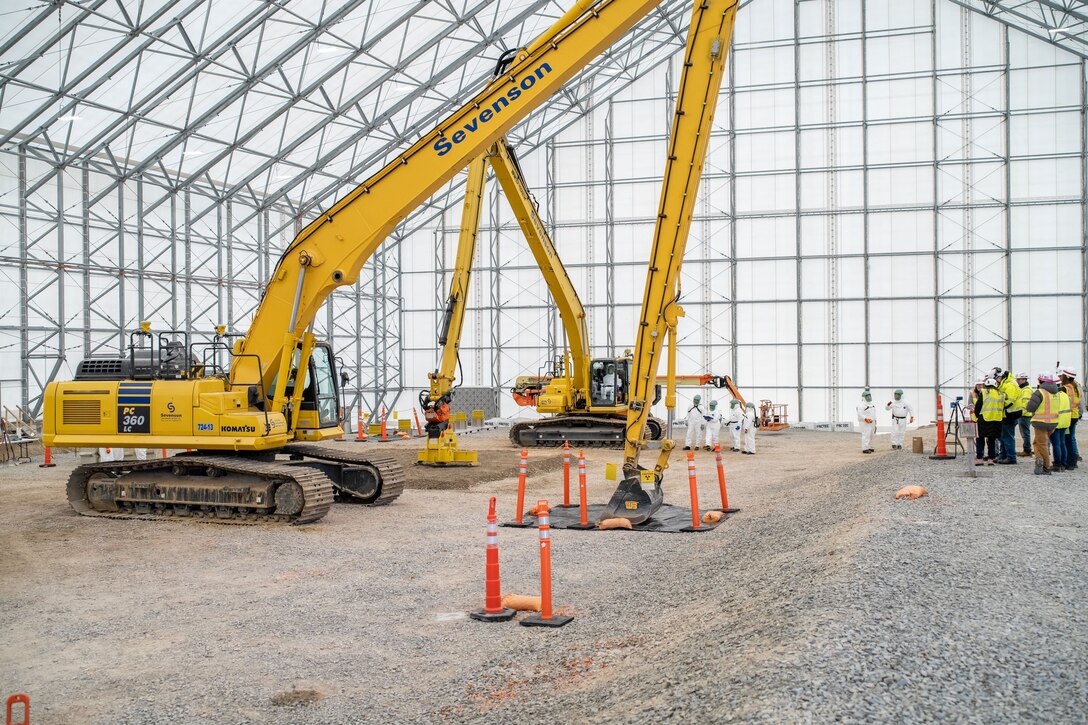 Two large yellow excavators are parked on the dirt floor inside a massive, white, tented, hangar-like structure. In the background, a group of workers in hard hats and protective suits observe.