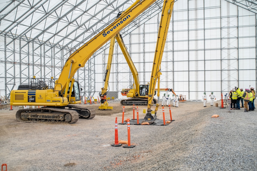 Two large yellow excavators are parked on the dirt floor inside a massive, white, tented, hangar-like structure. In the background, a group of workers in hard hats and protective suits observe.