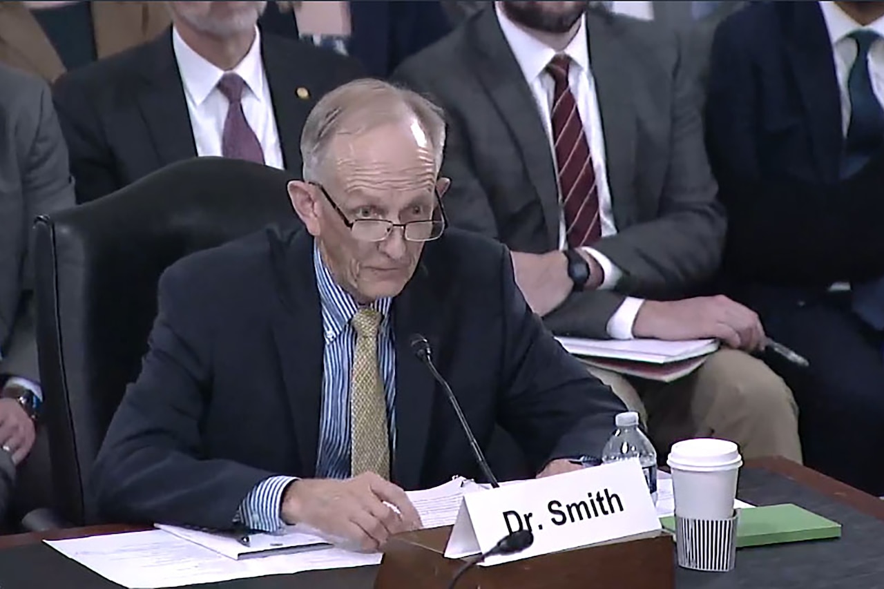 A man wearing a suit seated at a table speaks into a microphone.