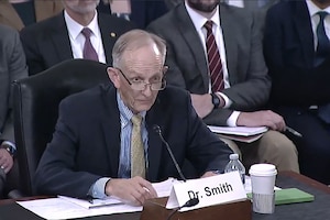A man wearing a suit seated at a table speaks into a microphone.