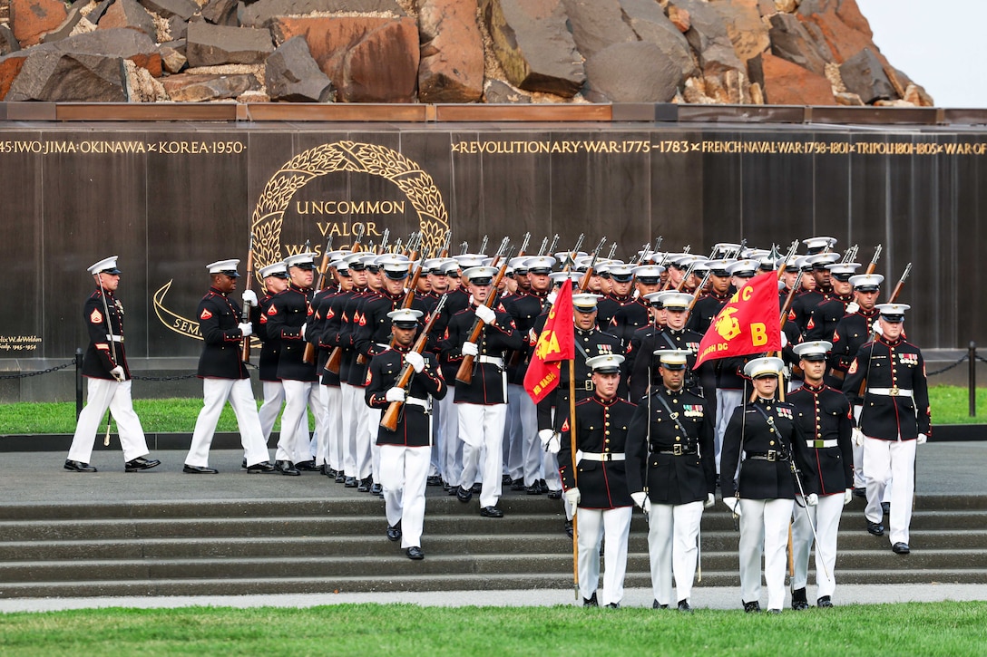 Dozens of Marines in ceremonial uniforms carry rifles while marching in formation down the steps of a memorial.