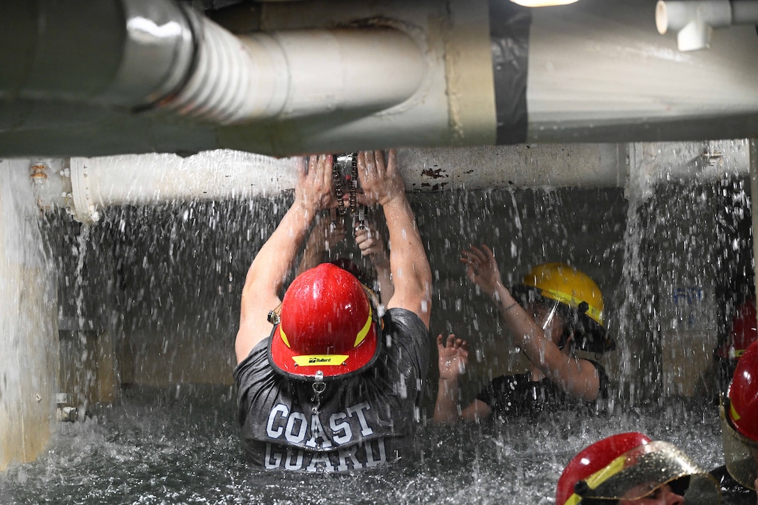Coast Guardsmen in red protective hats stand in flood water while trying to repair leaking pipes from above.