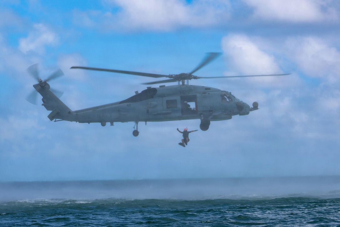 A sailor wearing wet gear jumps from a hovering helicopter into a body of water during the day.