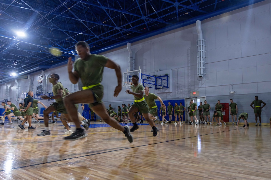 Marines wearing green athletic wear and reflective neon belts run full speed across a dimly lit basketball court.