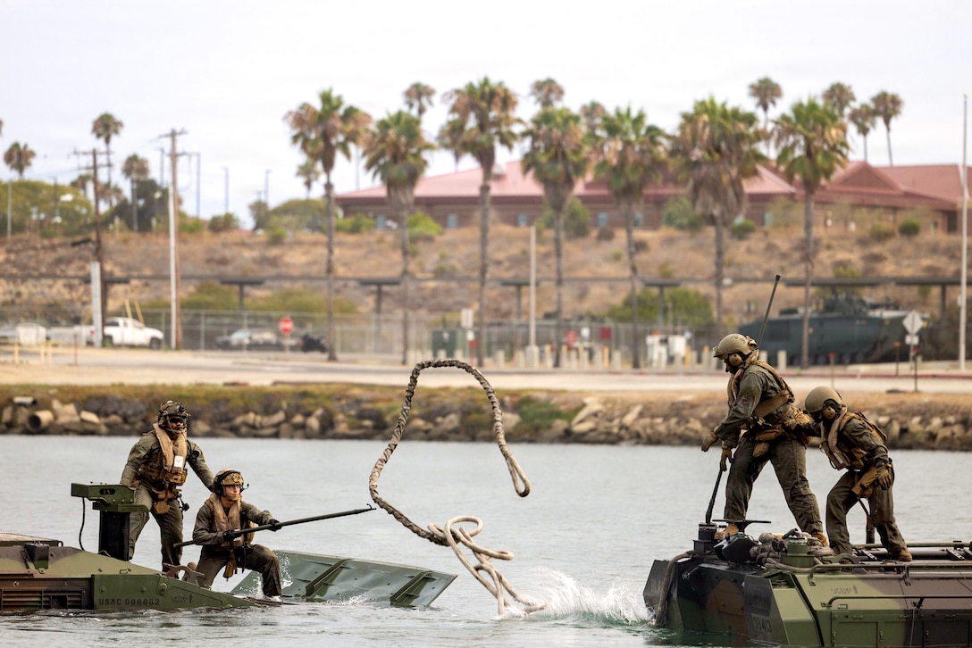 Two Marines aboard an amphibious vehicle throw a rope to two other Marines aboard another amphibious vehicle in a body of water during daytime.