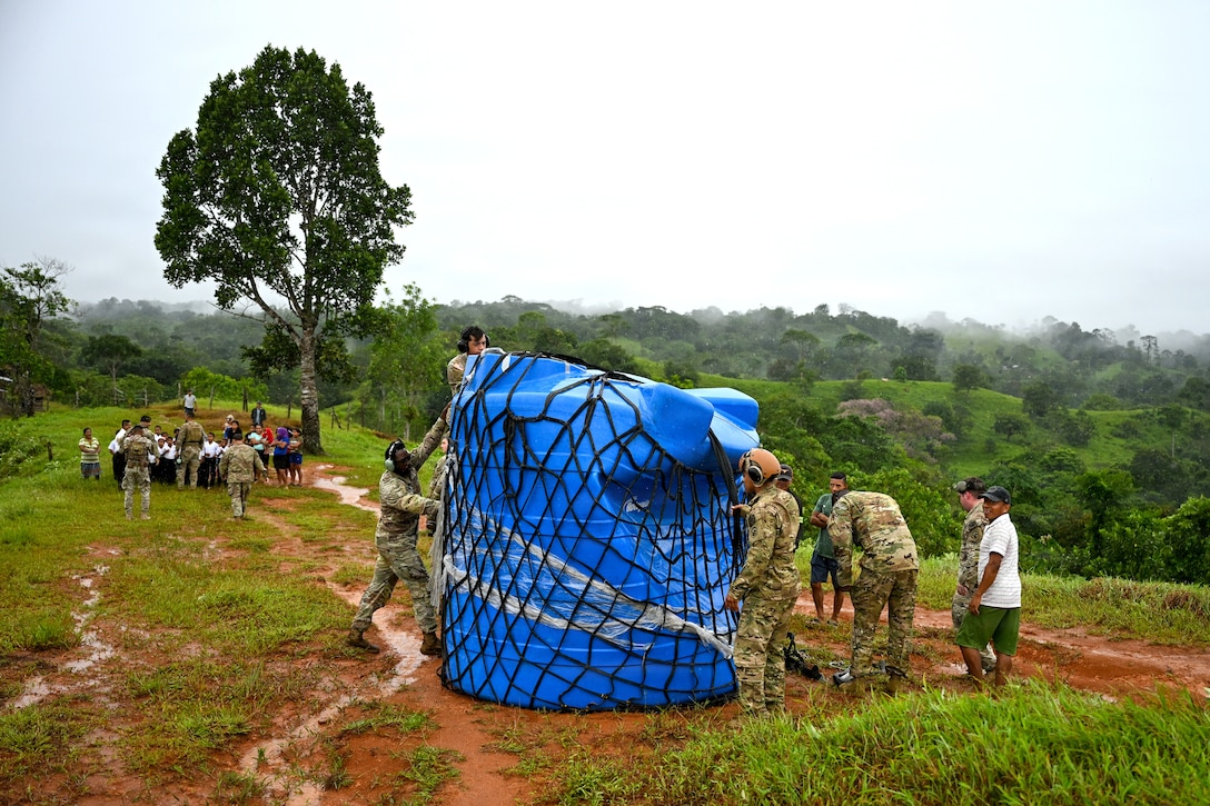 A group of about a half dozen service members in camouflage uniforms remove a net from a large blue water bladder while people in civilian clothes look on from a distance during daytime in a rural location with subtropical foliage.