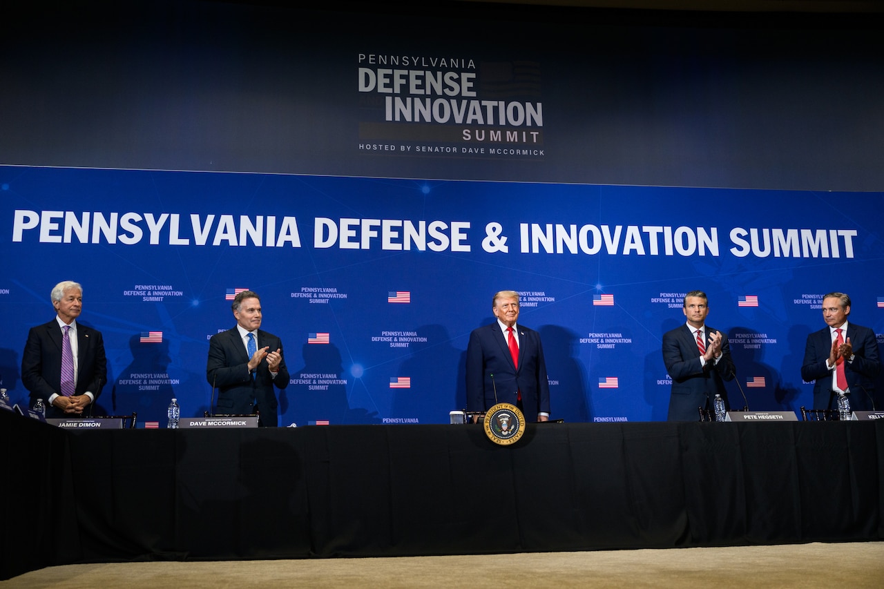 President Donald J. Trump stands with defense industry leaders against a blue background.
