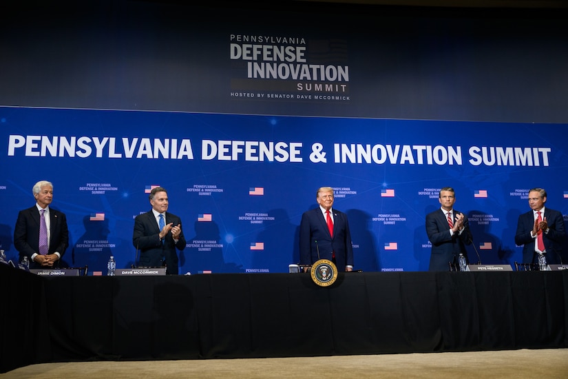 President Donald J. Trump stands with defense industry leaders against a blue background.