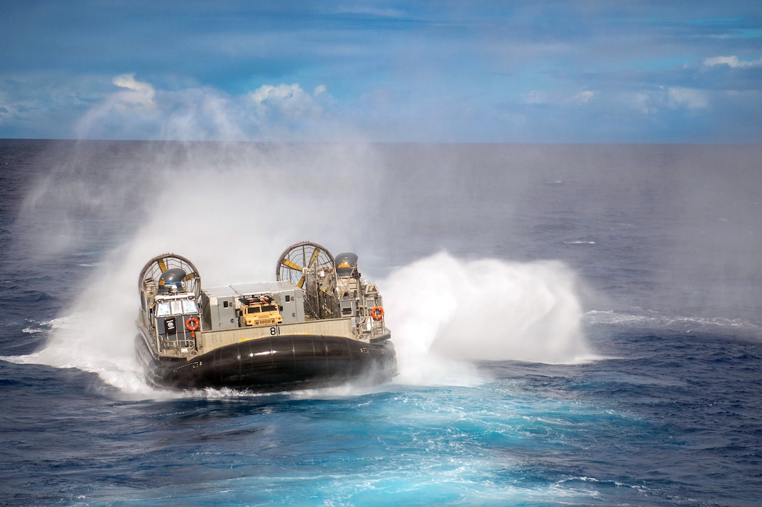 An air-cushioned landing craft sends up spray and makes waves on a body of water with blue water and a blue sky during daytime.