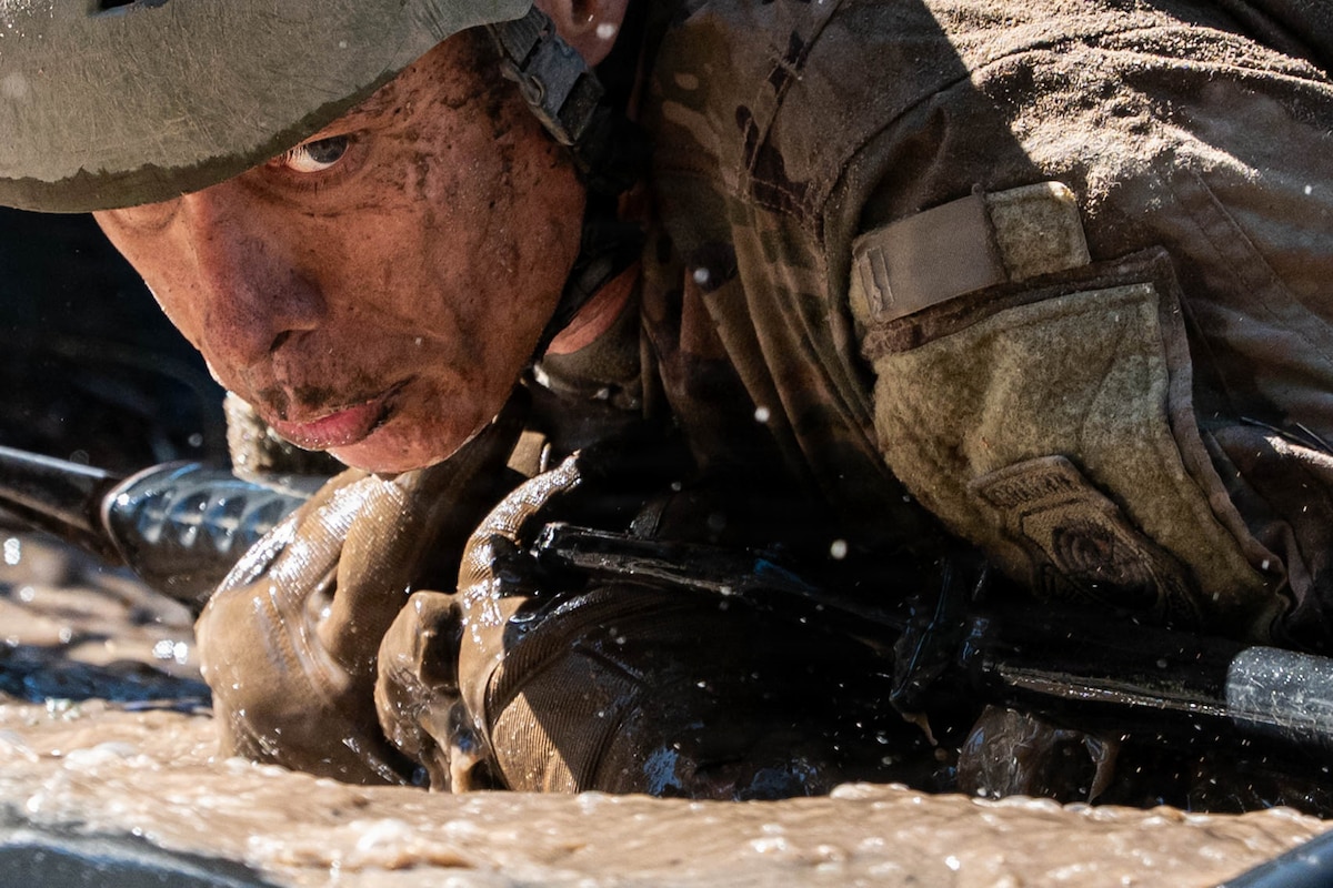 A service member wearing a camouflage military uniform and helmet crawls through mud carrying a rifle during daytime.