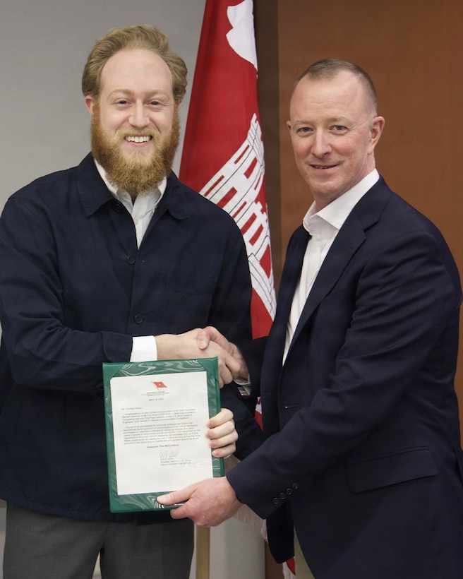 Two men are standing indoors, shaking hands in front of a red and white flag. The man on the left has a full beard and is smiling broadly. He is wearing a dark blue jacket over a light-colored collared shirt. He is holding a green document holder with a certificate inside. The man on the right has short hair and is also smiling. He is wearing a dark suit jacket over a white collared shirt.
