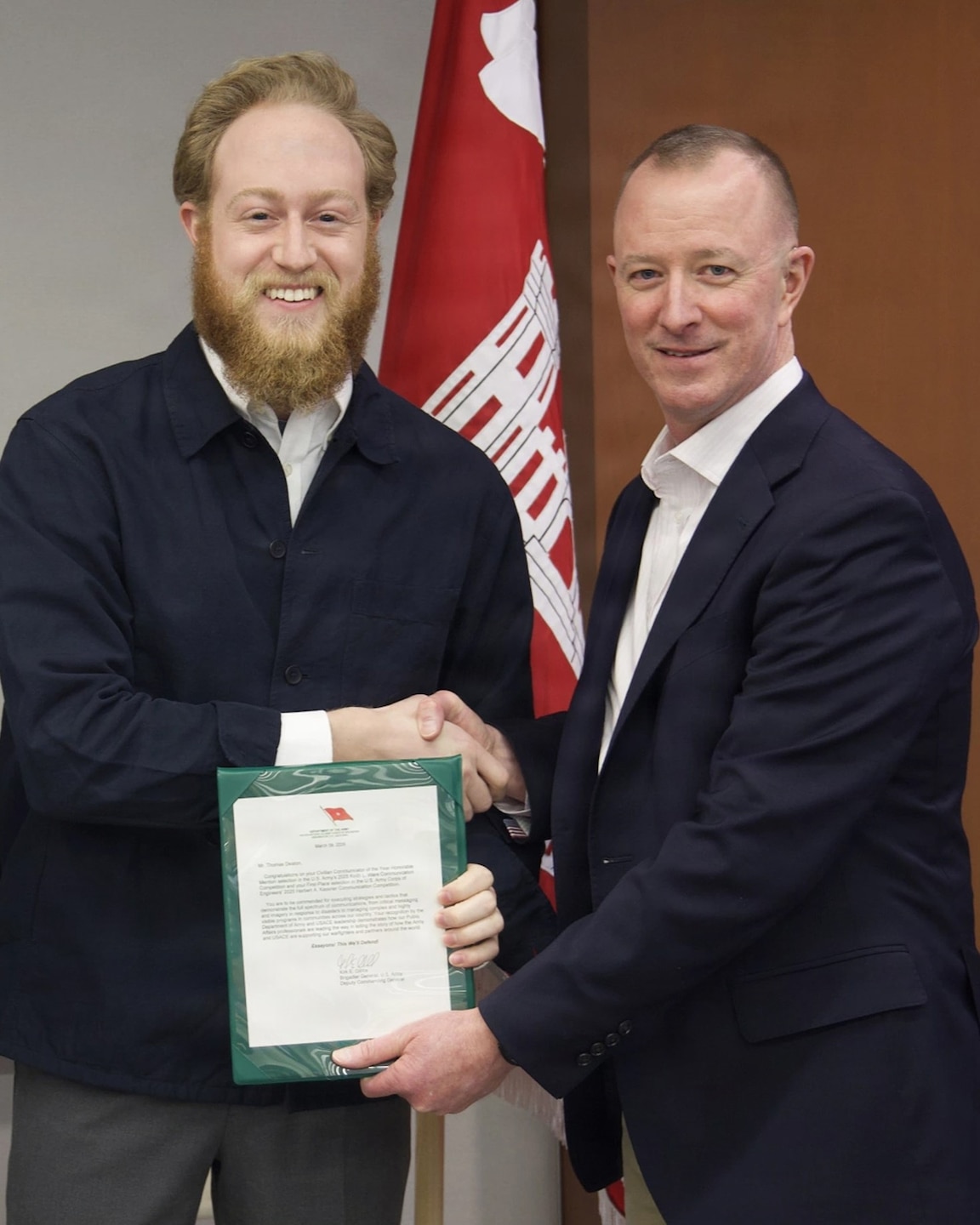 Two men are standing indoors, shaking hands in front of a red and white flag. The man on the left has a full beard and is smiling broadly. He is wearing a dark blue jacket over a light-colored collared shirt. He is holding a green document holder with a certificate inside. The man on the right has short hair and is also smiling. He is wearing a dark suit jacket over a white collared shirt.