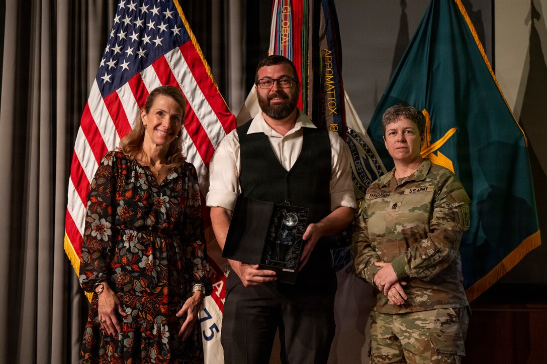 Three people are posing for a photo, standing in front of an American flag and other military-style banners. On the left, a woman with long hair smiles while wearing a dark, floral-patterned dress. In the center, a man with a beard and glasses, dressed in a white shirt and dark vest, holds a black box containing a glass award. On the right stands a U.S. Army soldier in a camouflage uniform with the name "FERGUSON" on her chest.