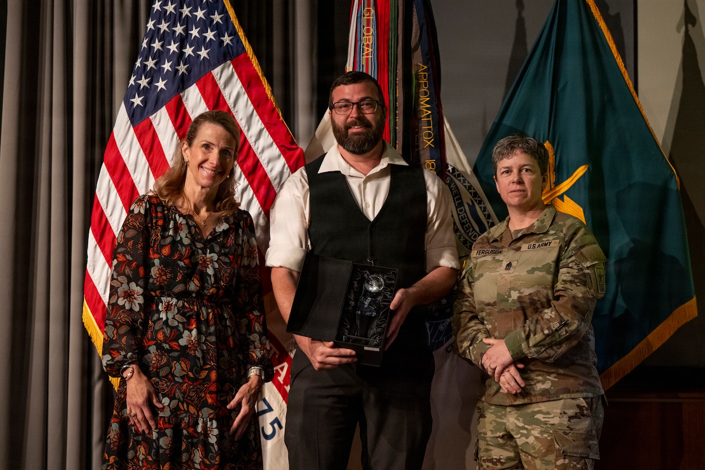Three people are posing for a photo, standing in front of an American flag and other military-style banners. On the left, a woman with long hair smiles while wearing a dark, floral-patterned dress. In the center, a man with a beard and glasses, dressed in a white shirt and dark vest, holds a black box containing a glass award. On the right stands a U.S. Army soldier in a camouflage uniform with the name "FERGUSON" on her chest.