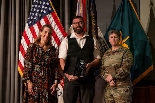 Three people are posing for a photo, standing in front of an American flag and other military-style banners. On the left, a woman with long hair smiles while wearing a dark, floral-patterned dress. In the center, a man with a beard and glasses, dressed in a white shirt and dark vest, holds a black box containing a glass award. On the right stands a U.S. Army soldier in a camouflage uniform with the name "FERGUSON" on her chest.