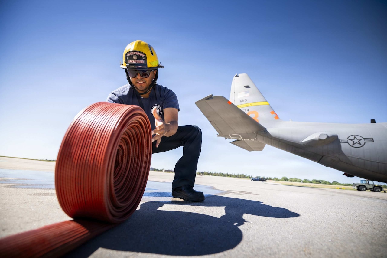 A firefighter rolls up a hose while kneeling on a tarmac. A plane and a forklift are in the background.