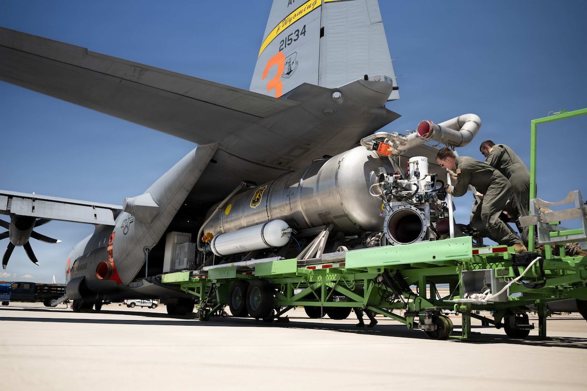 Two people dressed in flight suits push equipment into a plane while standing on a cargo loader.