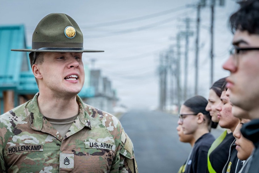 A man in a camouflage military uniform and large hat speaks to a group of people standing in formation.