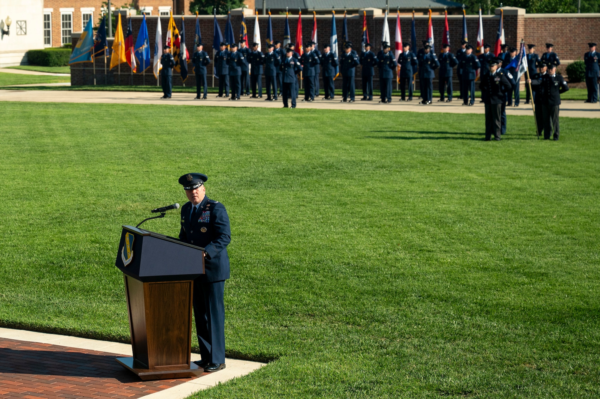 U.S. Air Force Col. stands behind a podium on a field in front of the U.S. Air and Space Force Honor Guard.
