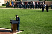 U.S. Air Force Col. stands behind a podium on a field in front of the U.S. Air and Space Force Honor Guard.