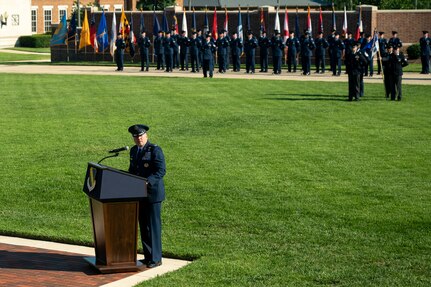 U.S. Air Force Col. stands behind a podium on a field in front of the U.S. Air and Space Force Honor Guard.