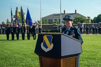 U.S. Air Force Col. stands behind a podium with The Chiefs Own symbol on a field in front of The U.S. Air and Space Force Honor Guard.