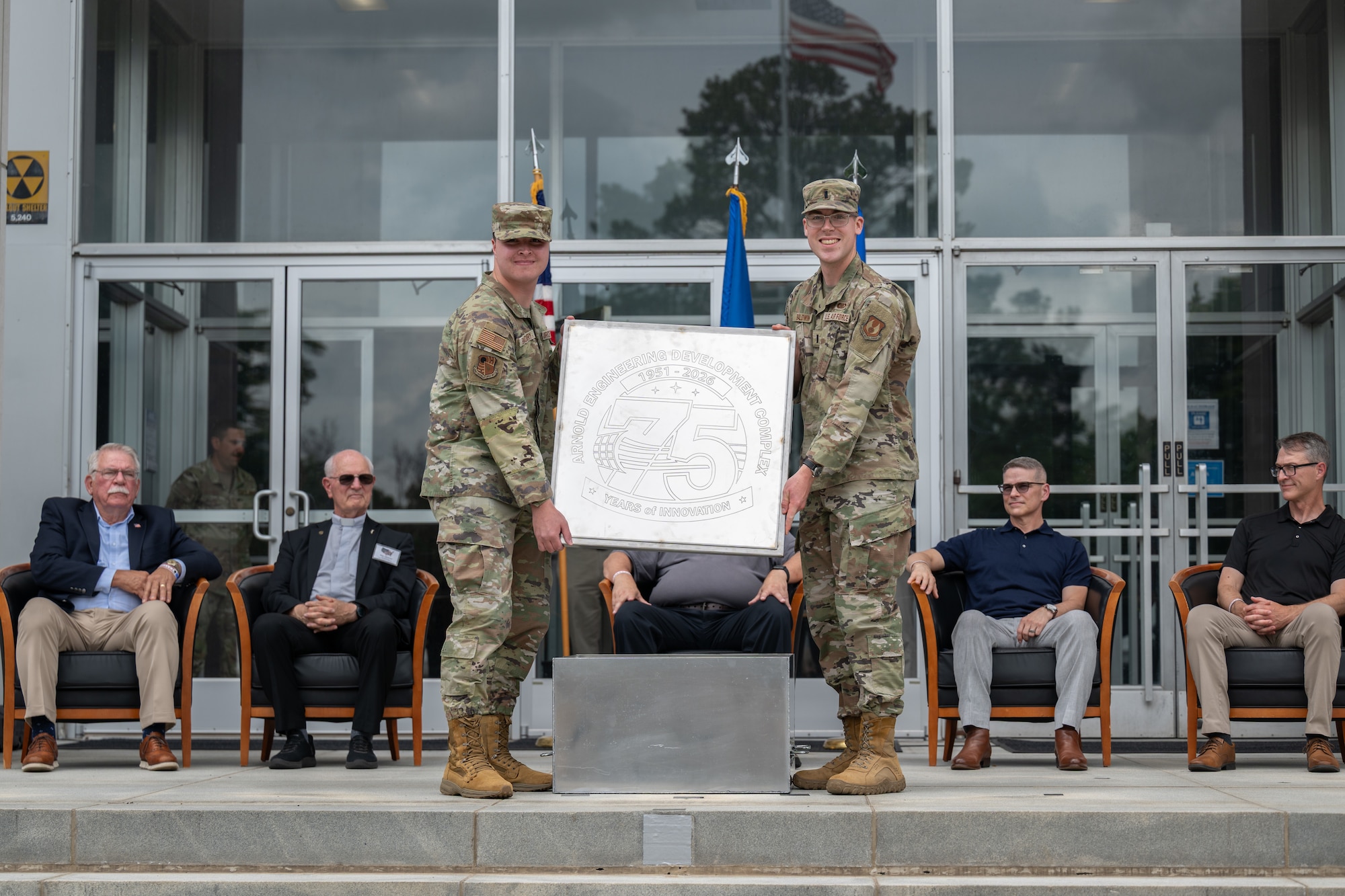 Senior Airman Christopher Bradbury, standing left, and 1st Lt. Thomas Baldwin, standing right, display the lid of the Arnold Engineering Development Complex time capsule unveiled June 27, 2026, during the AEDC 75th anniversary open house celebration at Arnold Air Force Base, Tenn., headquarters of AEDC. Former AEDC commander pictured seated, from left, are retired Air Force Col. Larry Graviss, retired Air Force Maj. Gen. Mike Wiedemer, retired Maj. Gen. Michael Brewer, retired Col. Raymond Toth and Maj. Gen. Scott Cain. (U.S. Air Force photo by Keith Thornburgh)
