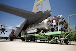 Two people dressed in flight suits push equipment into a plane while standing on a cargo loader.