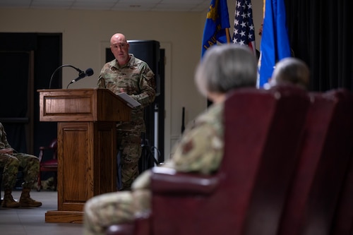 Col. Eric Bissonette speaks at a podium while looking at the incoming and outgoing Laughlin Medical Group commanders sitting in chairs.