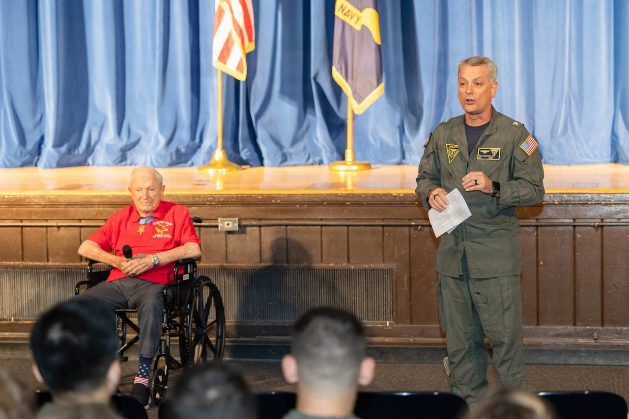 A man in a flight suit speaks to a group of people sitting in an auditorium while an elderly man in a wheelchair holds a microphone next to him.