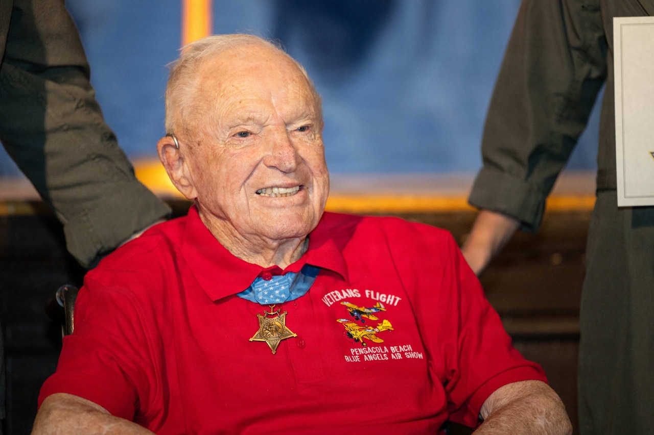 An elderly man in a wheelchair smiles while posing for a photo with a medal around his neck.