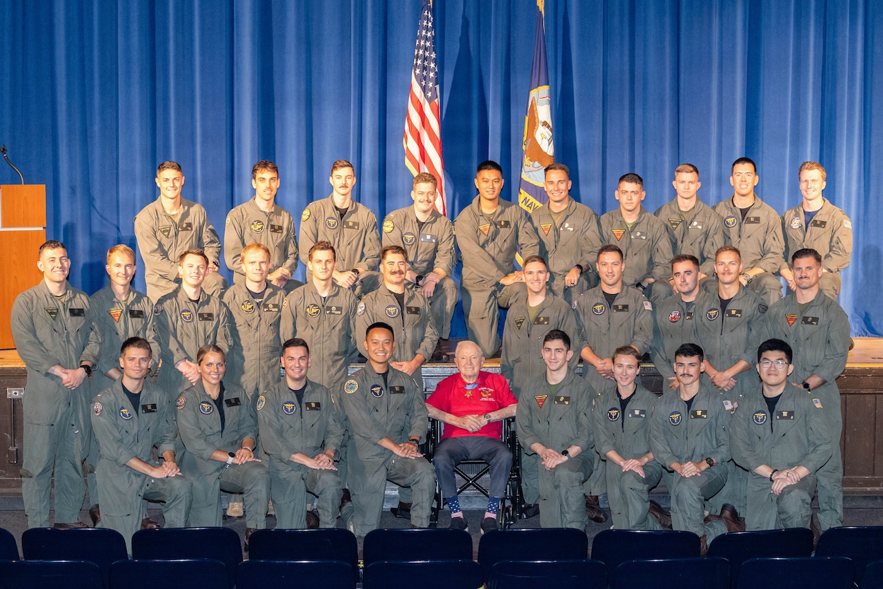 An elderly man in a wheelchair poses for a photo in an auditorium with dozens of men in flight suits.